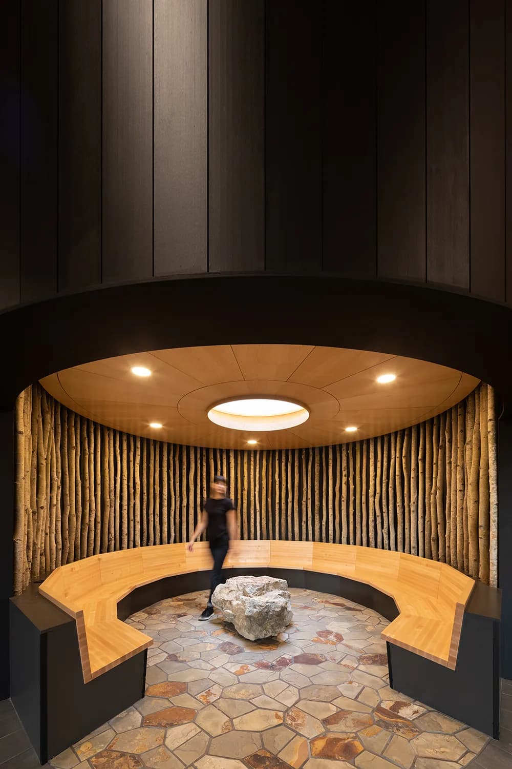 A cultural gathering room dressed with vertical local wood, with a curved timber bench and stone floor set around a central boulder. Ready for smudging.